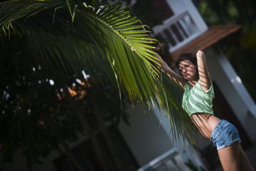 Young beautiful girl in shorts posing with a palm leaf in the summer on the background of the cottage. Palm tree casts shadows on the girl's face