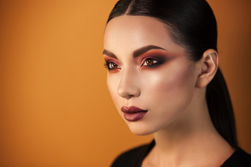 Fototapeta premium a Eurasian girl with sleek hair staring into the distance on an orange background, graceful make-up gleams her eyes and sensual lips.