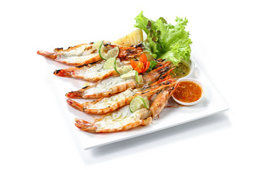Roasted cleaved black tiger prawns with boiled potatoes, slice of limes, fresh vegetables and chilli sauce on white square plate, isolated on white background with shadow, high angle front side view.