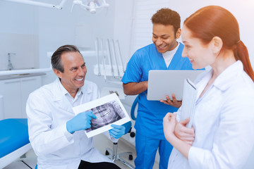 Funny moment. Professional dentist keeping smile on his face and sitting in his cabinet while holding picture in both hands