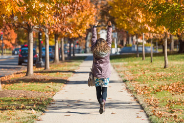 Young happy one woman, arms raised on sidewalk street walking in Washington DC, USA in alley of golden orange yellow foliage autumn fall trees