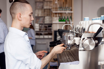 A young Barista guy with a stylish hairstyle works in a cafe and unscrews the filter holder from the coffee machine - 208314836