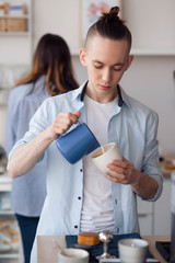 The Barista with stylish hairstyle stands and cooks cappuchino at work. He pours milk in coffee. - 208314854