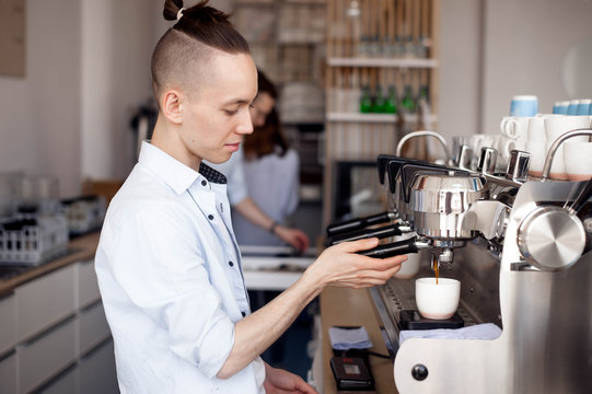 A young men Barista with a stylish hairstyle works in a cafe and brews coffee in the coffee machine holding the handle of the filter holder. Coffee flows into the Cup