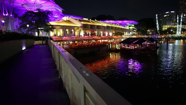 POV walking on the river in Clarke Quay Riverside area with cruise docked at pier in Singapore, Southeast Asia. Waterfront skyline reflected on Singapore River. Popular attraction for nightlife.