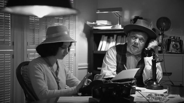 Scene tracks around 1940s newsroom with a reporter talking on candlestick phone while the other types on vintage manual typewriter.