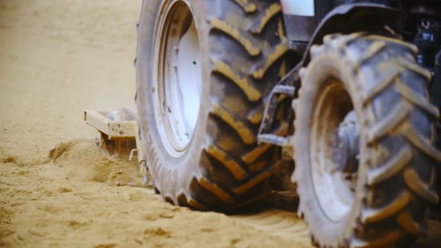 Tractor plow through sand close-up slow motion 4K. Long shot tracking tractor plows in focus while it loosens the sand of riding arena. Big tractor tires.