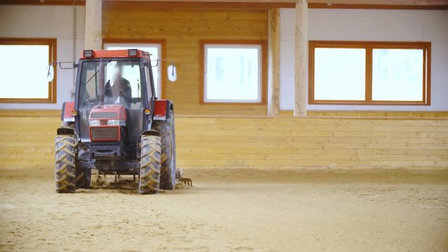 Tractor in riding arena plow through sand in slow motion 4K. Long shot wide view of farm tractor plowing through sand in horse riding arena. Vehicle driving towards the camera.