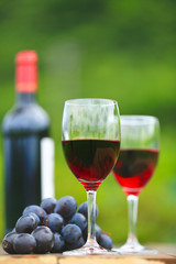 Two glasses Red Wine with wine bottle and fresh grapes on wooden table, defocused green outdoors background 1