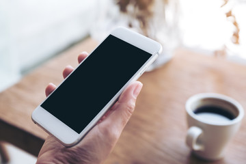 Mockup image of hand holding white mobile phone with blank black desktop screen in cafe