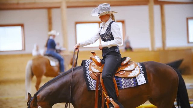 Attractive female western rider at a horsemanship competition 4K. Long shot side view of cowgirl in focus riding quarter horse inside the riding arena. More riders in the background out of focus.