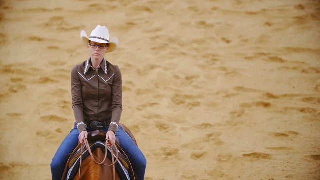 Cowgirl riding horse top view slow motion 4K. Extreme long shot tracking attractive female rider in focus riding horse in a straight line inside riding arena.