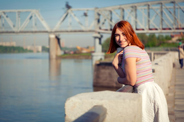 A young red-haired woman in a white romantic skirt, a pink top, drinks coffee and looks at the river on the city waterfront on a summer day