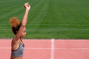 Sports exercises and stretching or preparing a runner to start at the stadium. A young beautiful African-American girl in a gray T-shirt raised her hands up, on a treadmill next to the green grass.