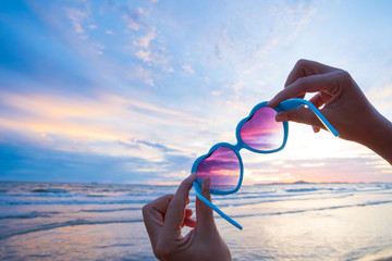 Soft focus on woman hand holding sunglasses in heart shape over sea and Sandy beach in background...