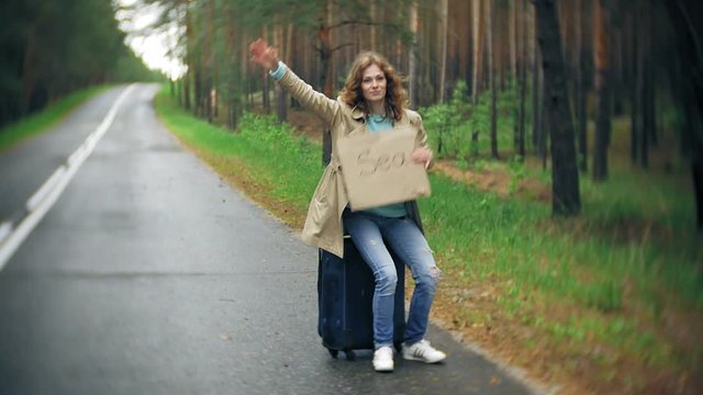 Young beautiful woman hitchhiking standing on the road with a suitcase on a table with an inscription SEA