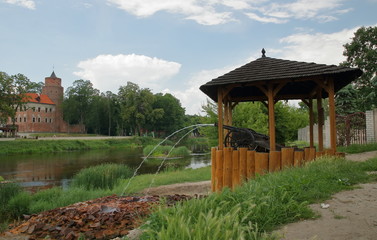 Widok na rzekę Wartę w Uniejowie, Polska, zamek na jej brzegu, otoczony parkiem, na pierwszm planie drewniana altana z dwoma armatkami, z których wytryskują dwa strumienie z uzdrowiskową wodą - obrazy, fototapety, plakaty