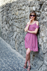 Graceful stylish young woman in red dress and sunglasses gladly posing while walk outside in good mood, street style. Outdoor close-up photo pleased girl in trendy summer dress, 