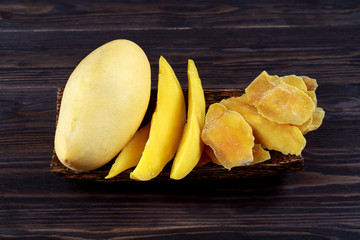 Dehydrated mango slices and fresh raw mango on wooden plate on dark background.