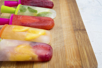 Homemade colorful fruity popsicle on a wooden board and against a white stone background. Strawberry, Lemon, Lemon with mint, Orange, Cherry, Multifruit
