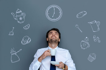 New cafe. Positive handsome elegant man holding a cup of coffee and smiling while looking at the clock above his head