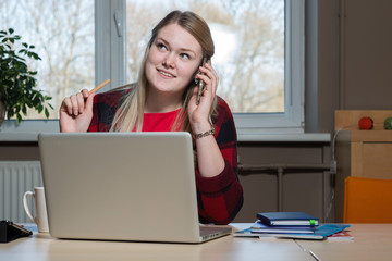 Obraz premium A smiling blonde woman sitting at a laptop and talking over a cell phone.