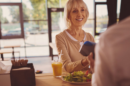 Satisfied customer. Charming cheerful senior woman giving a bank card to barista and paying for her salad and juice in cafe while smiling - Powered by Adobe
