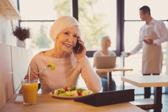 Pleasant conversation. Charming elderly woman having a conversation on the phone with her friend while eating salad in cafe and reading from her tablet