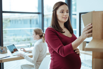 Need this one. Charming pregnant female standing in semi position and keeping smile on face while being in the office
