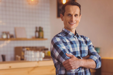 Obraz premium Upbeat owner. Cheerful young male cafe owner in a checked shirt folding his arms across his chest and smiling while posing for the camera in his own cafe