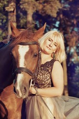 Obraz premium Portrait of a beautiful blonde woman in a chic dress with a horse walking in a pine forest in a stable