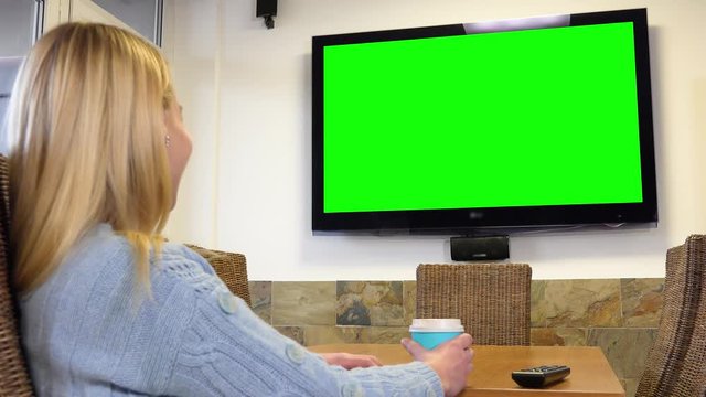 A woman sits at a table in a living room, watches a TV with a green screen and laughs