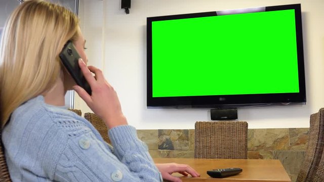 A woman sits at a table in a living room and talks on a smartphone - a TV with a green screen in the background