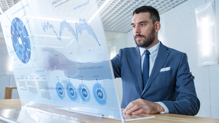 In the Near Future Successful Businessman Works on Computer with Transparent Display that Shows Stock Market Ticker Data, Numbers, Rises and Downs.