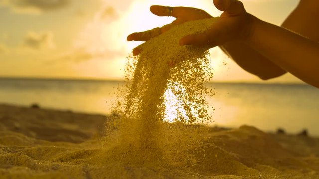 SLOW MOTION, CLOSE UP: Young woman on summer holiday letting coarse sand slip through her fingers. Unrecognizable girl lifts up a handful of white sand over the sunset shining on the exotic beach.
