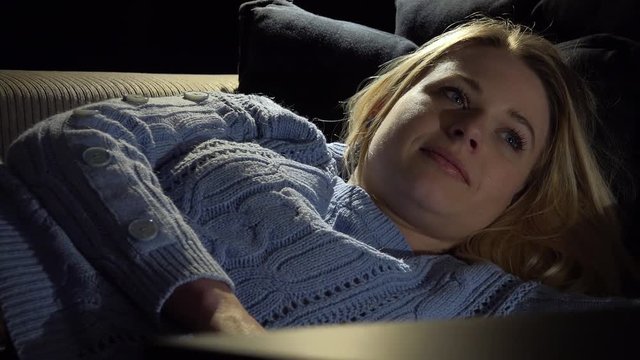 A young beautiful woman lies on a couch in a dark room and watches a TV - closeup on the woman