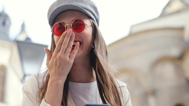 happy fashionable brunette woman laughing while using modern smart phone mobile on the street looking at screen stylish accessories pretty lady smiling joy fun happiness trendy red sunglasses portrait