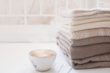 Stack of warm knitwears and cup of hot tea on windowsill on white window background.