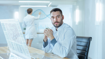 Obraz premium In the Futuristic Laboratory Male Scientists Looking into Camera. He is Working on the Transparent Display Computer. In the Background Female Scientists Writing on Blackboard Sequenced Human DNA.