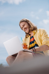 low angle view of smiling young man holding cocktail and using laptop at beach