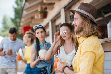 side view of smiling young man looking away while drinking cocktails with friends at beach bar