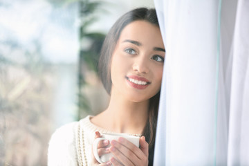 Beautiful young woman with cup of hot tea at home, view through window