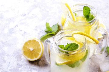 Two mason jar glasses of homemade refreshing lemonade with slices of organic ripe lemon, whole and halved, mint, ice cube on grunged grey concrete table background. Close up, top view, copy space.