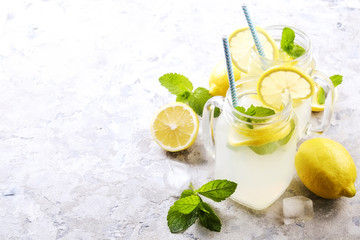 Two mason jar glasses of homemade refreshing lemonade with slices of organic ripe lemon, whole and halved, mint, ice cube on grunged grey concrete table background. Close up, top view, copy space.