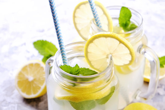 Two mason jar glasses of homemade refreshing lemonade with slices of organic ripe lemon, whole and halved, mint, ice cube on grunged grey concrete table background. Close up, top view, copy space.