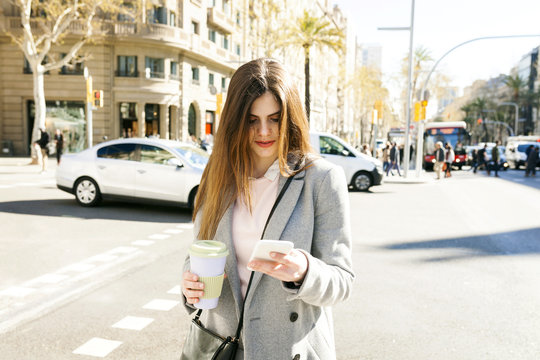 Spain, Barcelona, young woman with coffee to go standing at roadside looking at cell phone