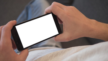 Mock up image of young man holding black mobile phone with blank white screen in landscape mode while sitting