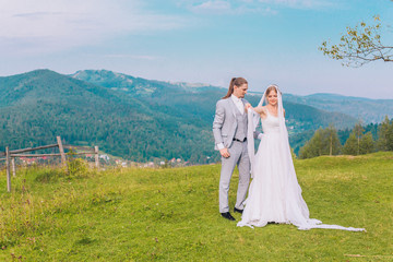 Handsome groom is gently holding his elegant new wife under pine tree. Mountain landscape. Young couple. Wedding day. 