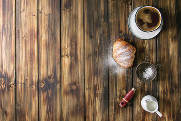 Fototapeta na wymiar Homemade croissant with sugar powder, cup of coffee, jug of milk, vintage sieve over wooden plank background. Flat lay, space.