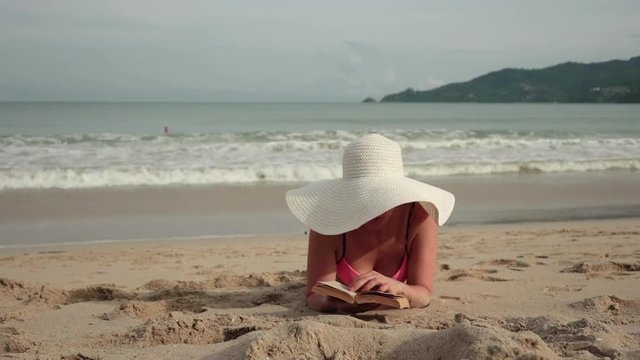 Young women in a bikini and sun hat reading a book while sunbathing at the beach